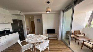 A modern kitchen with white cabinets and a black countertop, a dining area with a round table and white chairs, and a balcony with wooden furniture. - Luxury A/C Apartment Near Beach and Shops - 2 Rooms, Parking, High-Quality Amenities (La Grande-Motte)