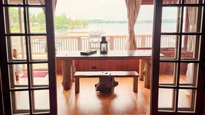 Un interior de madera con un banco y una mesa, ventanas amplias con cortinas y vista a un cuerpo de agua.