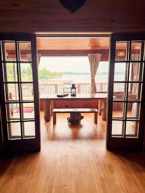 A wooden interior with a bench and a table, large windows with curtains, and a view of a body of water.