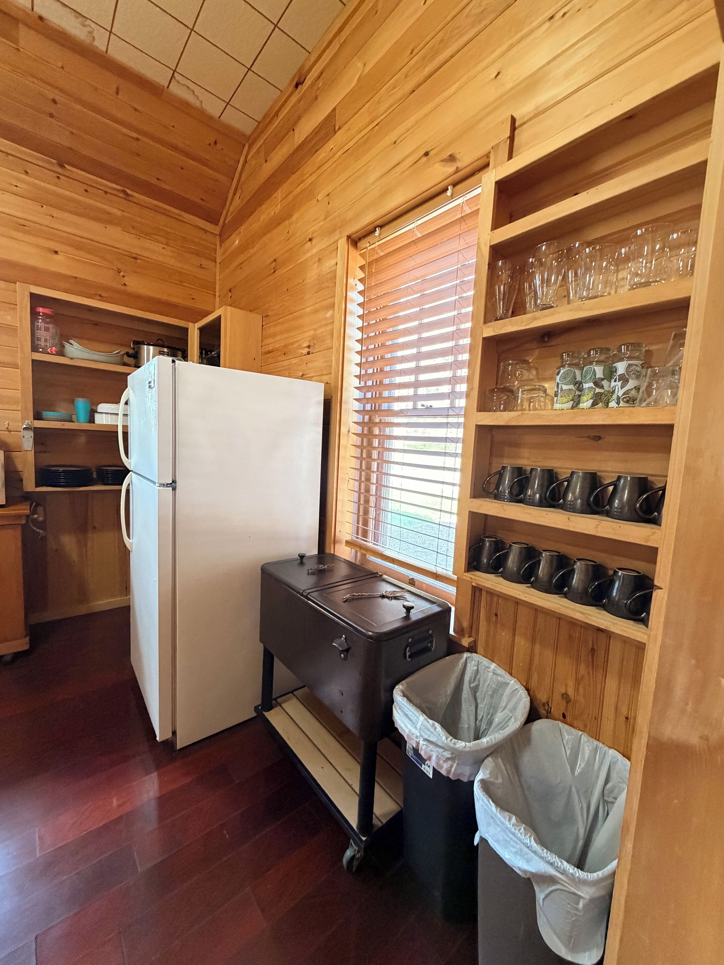 A compact kitchen with wooden walls, a white refrigerator, a small stove, and a trash bin.
