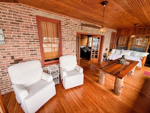 Una habitación acogedora con pared de ladrillo, techo de madera, sillas blancas tapizadas, una mesita de centro de madera y un banco de madera.