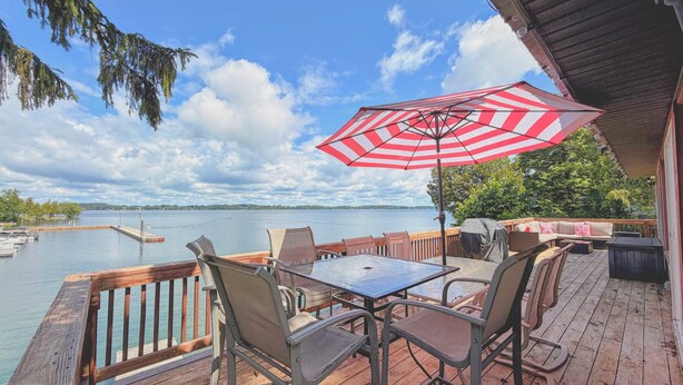 Una terraza de madera con mobiliario exterior, un paraguas a rayas rojas y blancas, y vista a un lago.