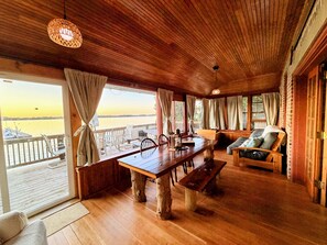 A wooden-floored room with a large table, chairs, and a sofa, overlooking a body of water.