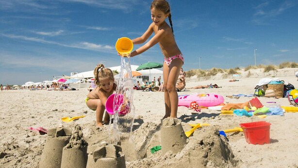 Due bambini giocano con la sabbia su una spiaggia, con ombrelloni e diversi giochi da spiaggia sullo sfondo.