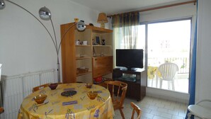 A dining area with a round table, chairs, a bookshelf, a television, and a window with a view. - Charming Studio by Beach with Private Parking and Garden Views (La Grande-Motte)
