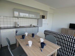 A compact kitchen with a dishwasher, a coffee maker, and a microwave. There is a dining table with four mugs and a spoon. A sofa with a patterned cover is in the background. - Charming 2-Room Apartment with Pool, Near Beach, Sleeps 5 (La Grande-Motte)