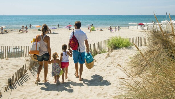 Ein Sandstrand mit einer Holzzahlung, Menschen die entlanggehen und Sonnenschirmen im Hintergrund.