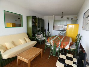 Un espacio habitable compacto con sofá, una pequeña mesa de madera, una zona de comedor con sillas, una cocina y un espejo en la pared.