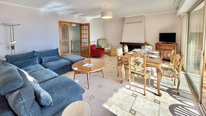 A living room with a blue sofa, a dining table with chairs, a TV, and a fireplace. - Terraced house, sea view - 6 guests (Ajaccio)