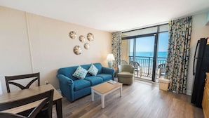 En stue med sofa, sofabord, spisebord og stole. Der er en balkon med havudsigt.