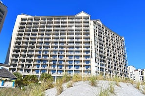 A multi-story hotel with a grid of balconies, located near a sandy area with sparse vegetation. - Amazing Oceanfront, Accessible Beach, Amazing View (North Myrtle Beach)