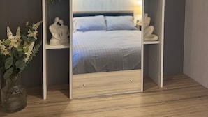 Een slaapkamer met een bed, nachtkastje, spiegel en planken met decoratieve objecten.