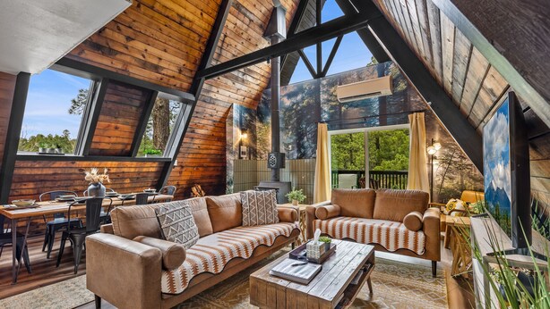 A cozy loft-style room with a wooden ceiling, large windows, a sofa, a coffee table, and a dining area. - The Needles A-Frame Cabin! Grill/Hot tub/Pet friendly! (Flagstaff)