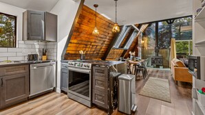 A modern kitchen with stainless steel appliances, a wooden accent wall, and a dining area with a view. - The Needles A-Frame Cabin! Grill/Hot tub/Pet friendly! (Flagstaff)