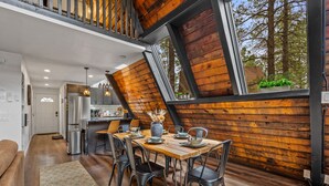 A modern kitchen with a large window, wooden walls, and a dining area with metal chairs. - The Needles A-Frame Cabin! Grill/Hot tub/Pet friendly! (Flagstaff)