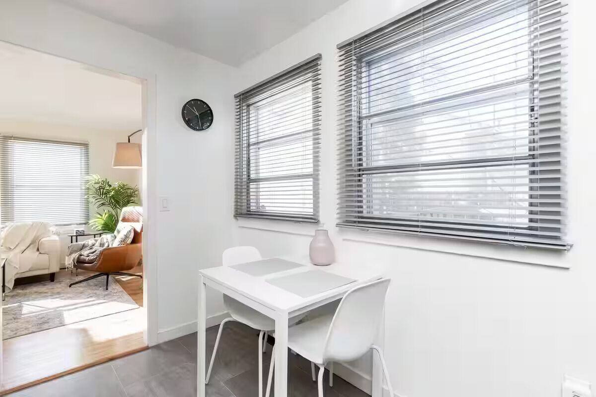 Una habitació moderna amb una taula blanca, dues cadires, un rellotge i grans finestres amb persianes.