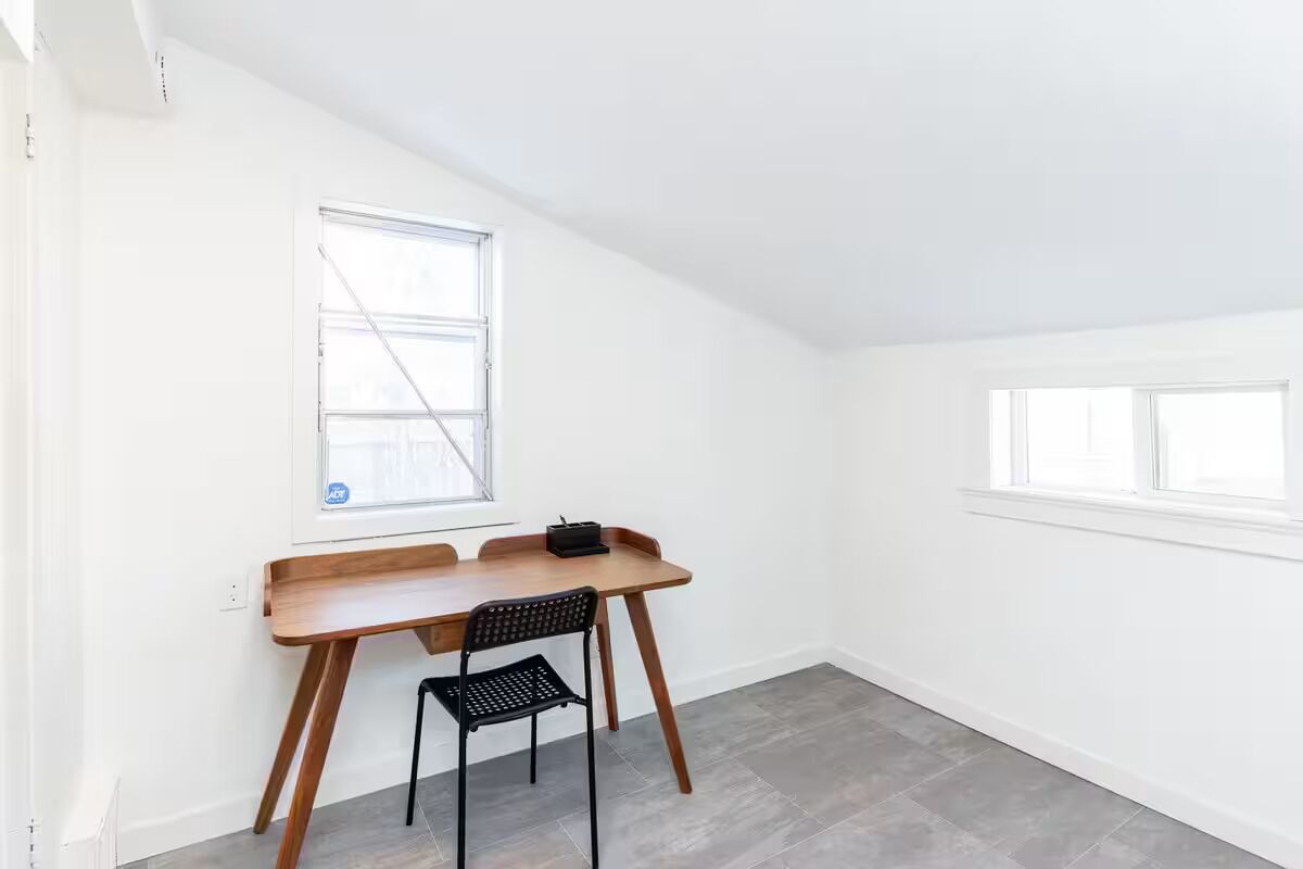 Une pièce avec un bureau en bois, une chaise noire et deux fenêtres.