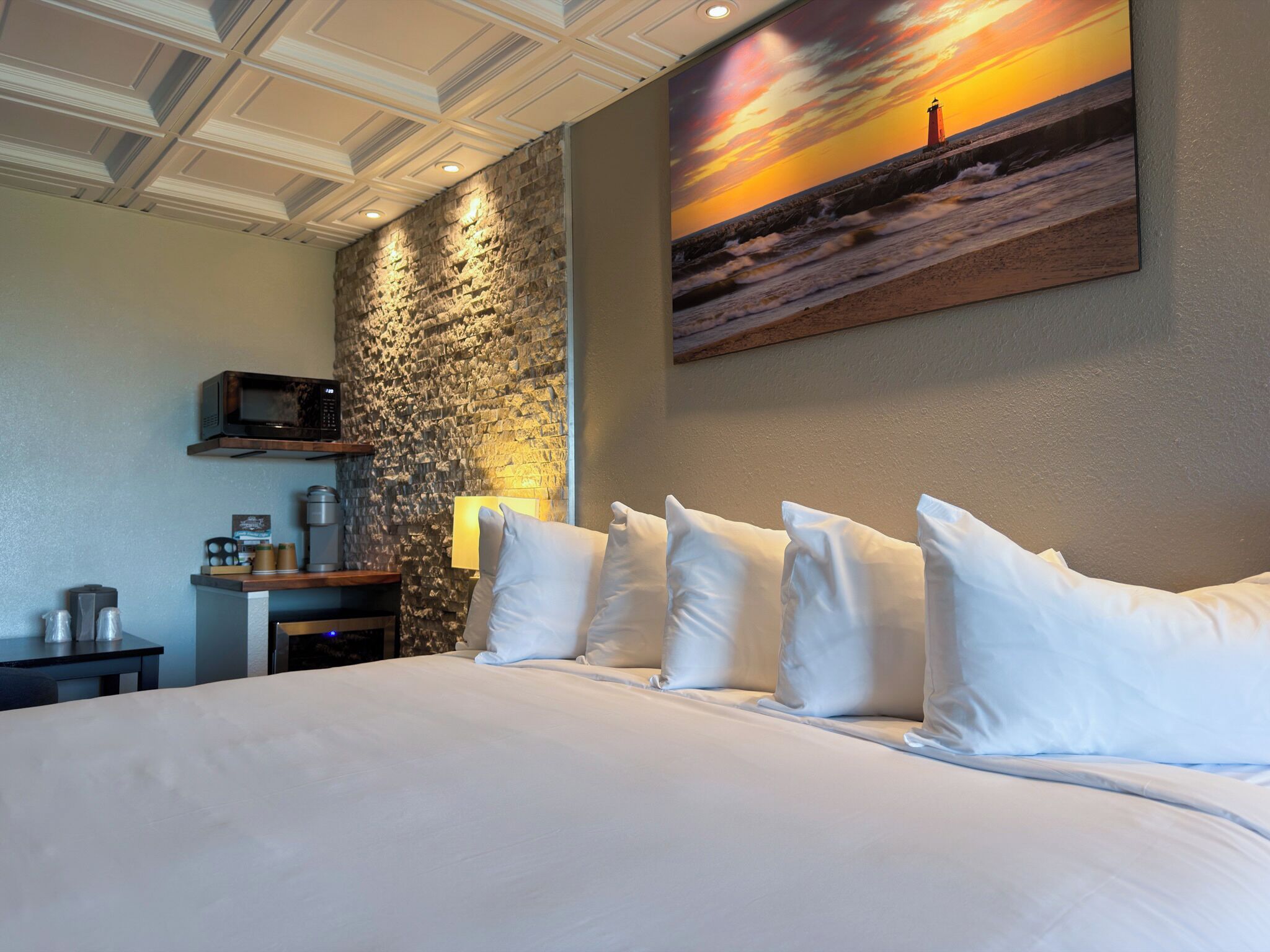 A hotel room with a large bed, a stone accent wall, a painting of a sunset, and a small desk with a lamp.