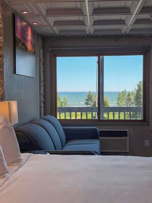 A bedroom with a large window, a bed, a blue sofa, a lamp, and a painting on the wall. - Manistique Sands (Manistique)
