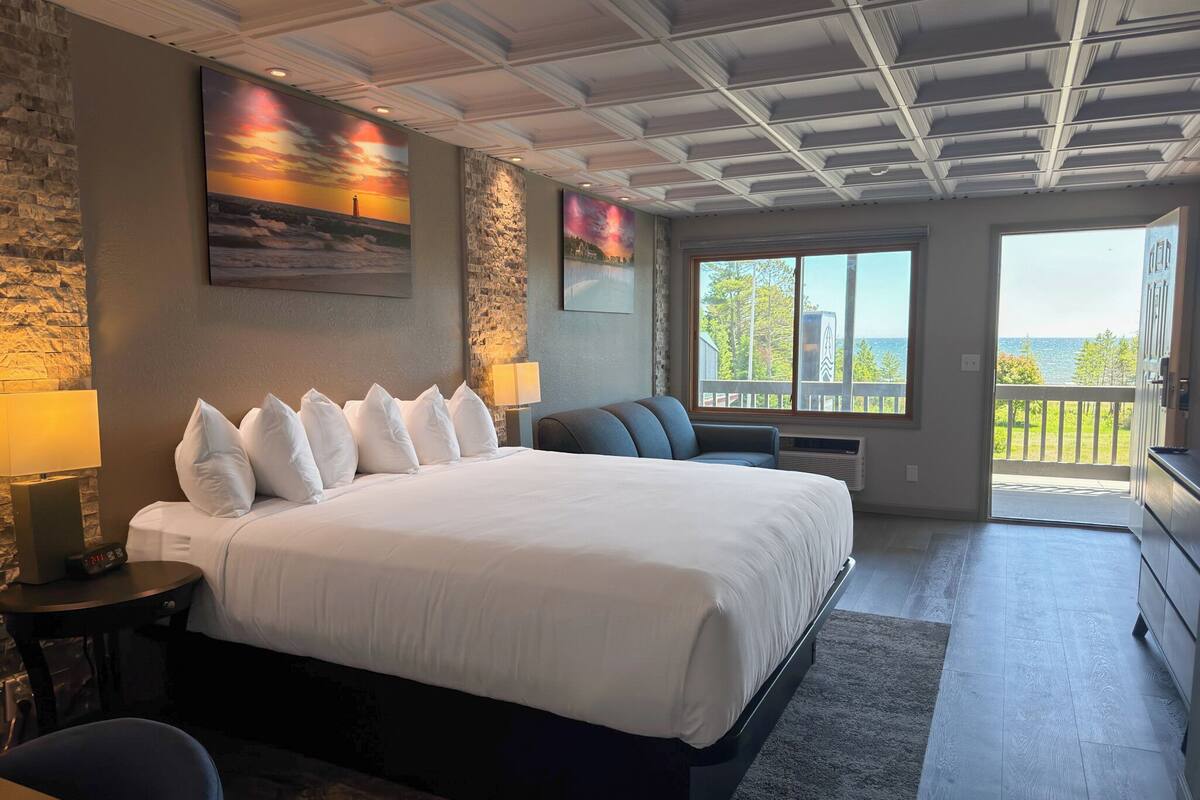 A hotel room with a large bed, a stone accent wall, a painting, a window with a view, and a balcony.