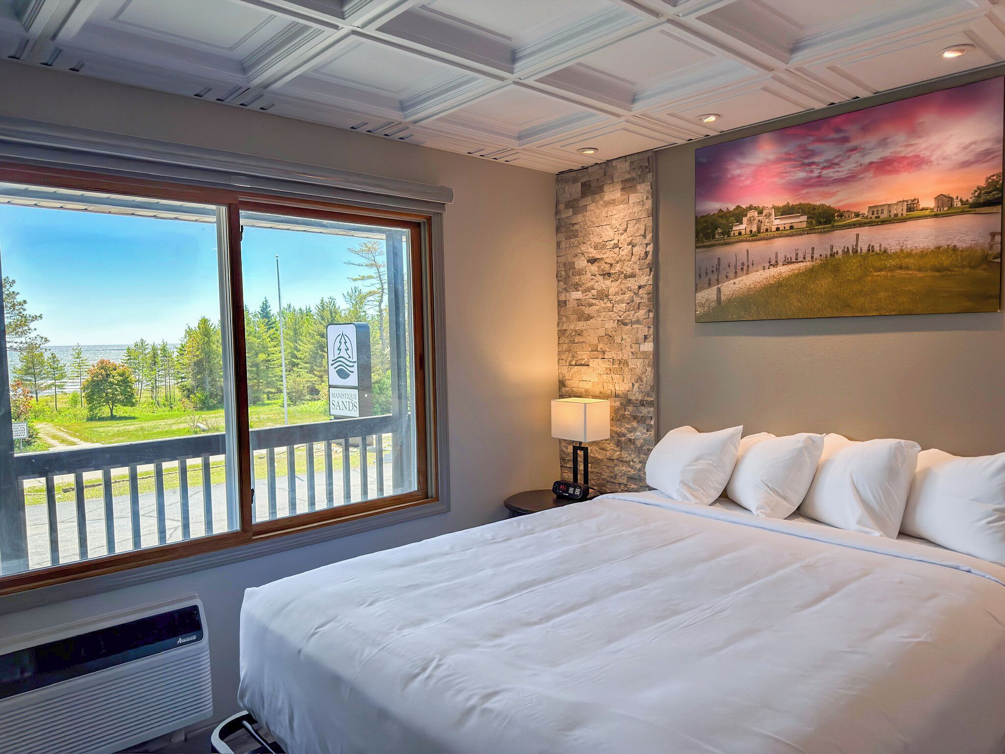 Hotelkamer met een groot bed, nachtkastje met lamp, stenen accentmuur, schilderij van een landschap en uitzicht op het omliggende landschap door een raam.