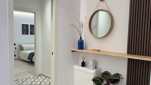 Un pasillo moderno con una puerta blanca, un espejo, un jarrón azul, una planta en maceta y una estantería con un objeto blanco.