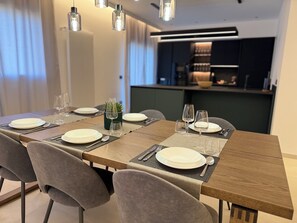 Zona pranzo con un lungo tavolo di legno apparecchiato per quattro persone, sedie rivestite in grigio e lampade a sospensione.