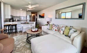 A modern living room with a white sofa, a coffee table, and a kitchen area with white cabinets and a dining table. - Puako Penthouse 409 ~ Top Floor Unit ~ Pool (Kamuela)
