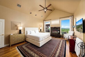 A bedroom with a bed, a ceiling fan, a nightstand, a dresser, a large window with a view, and a TV.