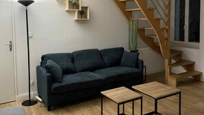 A modern living room with a dark-colored sofa, two small wooden tables, a floor lamp, a staircase, and a wall-mounted shelf with plants.