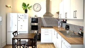 A compact kitchen with a refrigerator, double oven, wooden dining table, and white cabinets.
