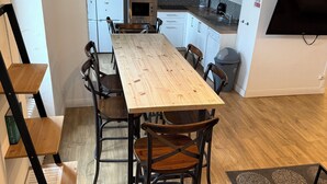 A modern kitchen with white cabinets, a wooden dining table, and a black TV mounted on the wall.