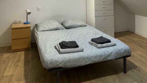 A bedroom with a sloped ceiling, a skylight, a bed with patterned bedding, a bedside table with a lamp, and a dresser.