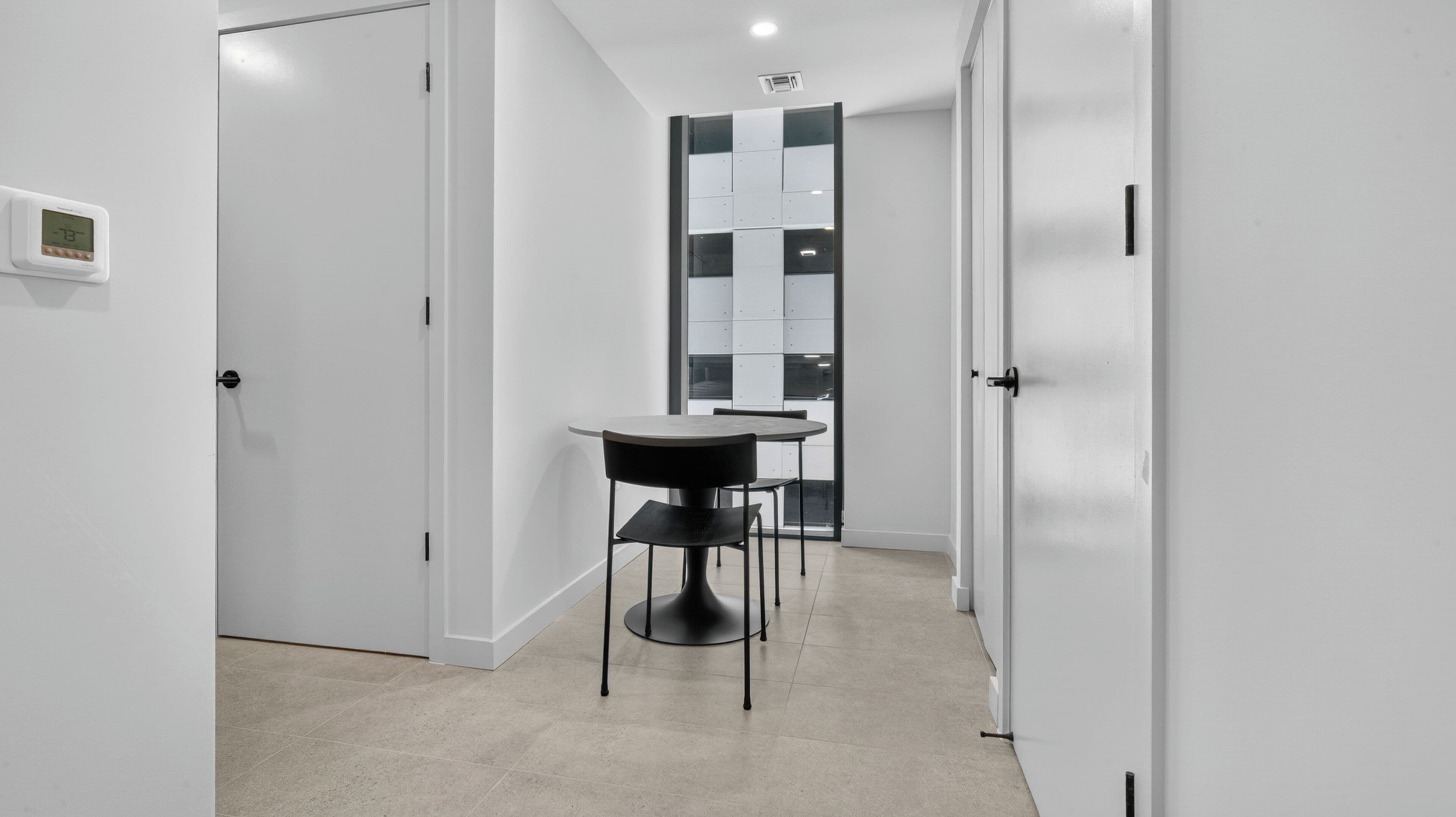 A hallway with a dining table and chairs, white walls, and a door with a thermostat.