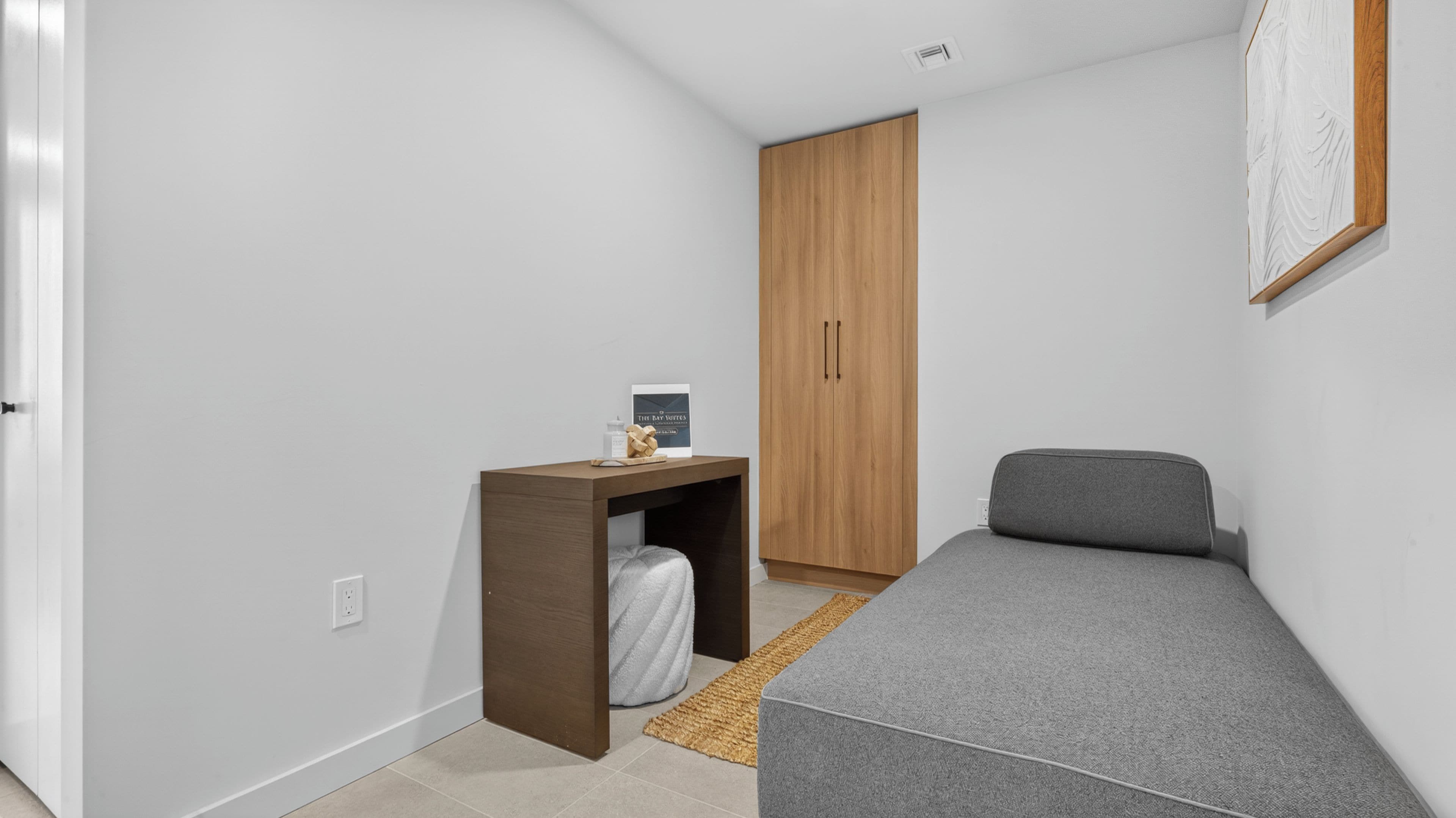 Une petite pièce comprenant un lit, une armoire en bois, un bureau avec une chaise et un tableau encadré accroché au mur.