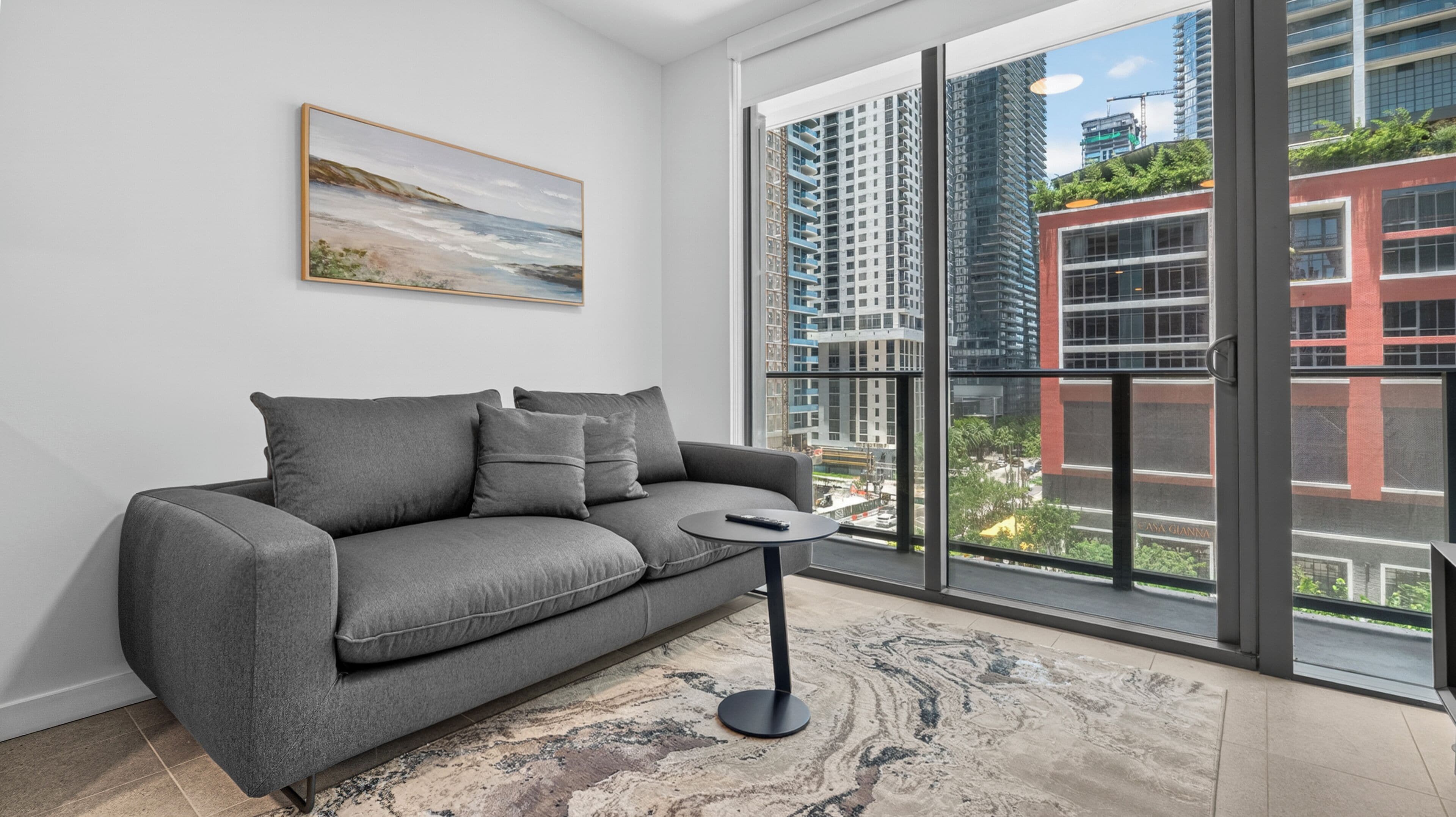 A modern living room with a grey sofa, a small round table, and a large window offering a view of high-rise buildings.