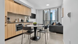 A modern kitchen with built-in appliances, a dining area with a round table and chairs, a grey sofa, and a large window with a city view. - The Urban Suites - 501 First Residence (Miami)