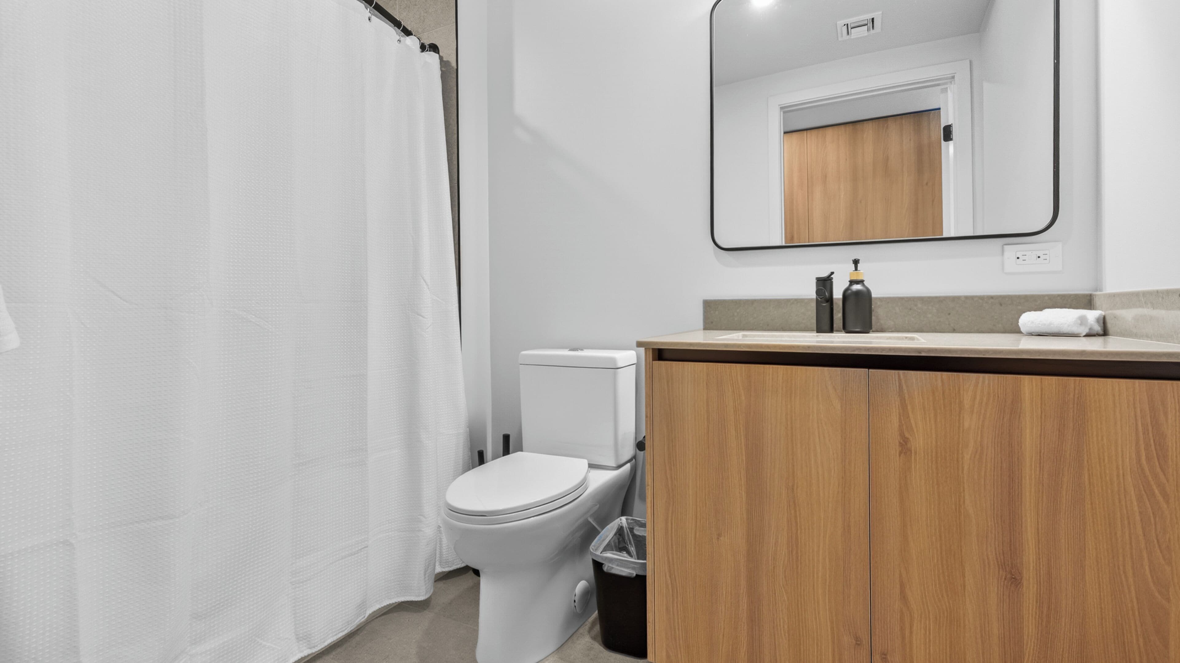 Un bagno moderno con il water bianco, un mobile bagno in legno, uno specchio e una tenda per la doccia.
