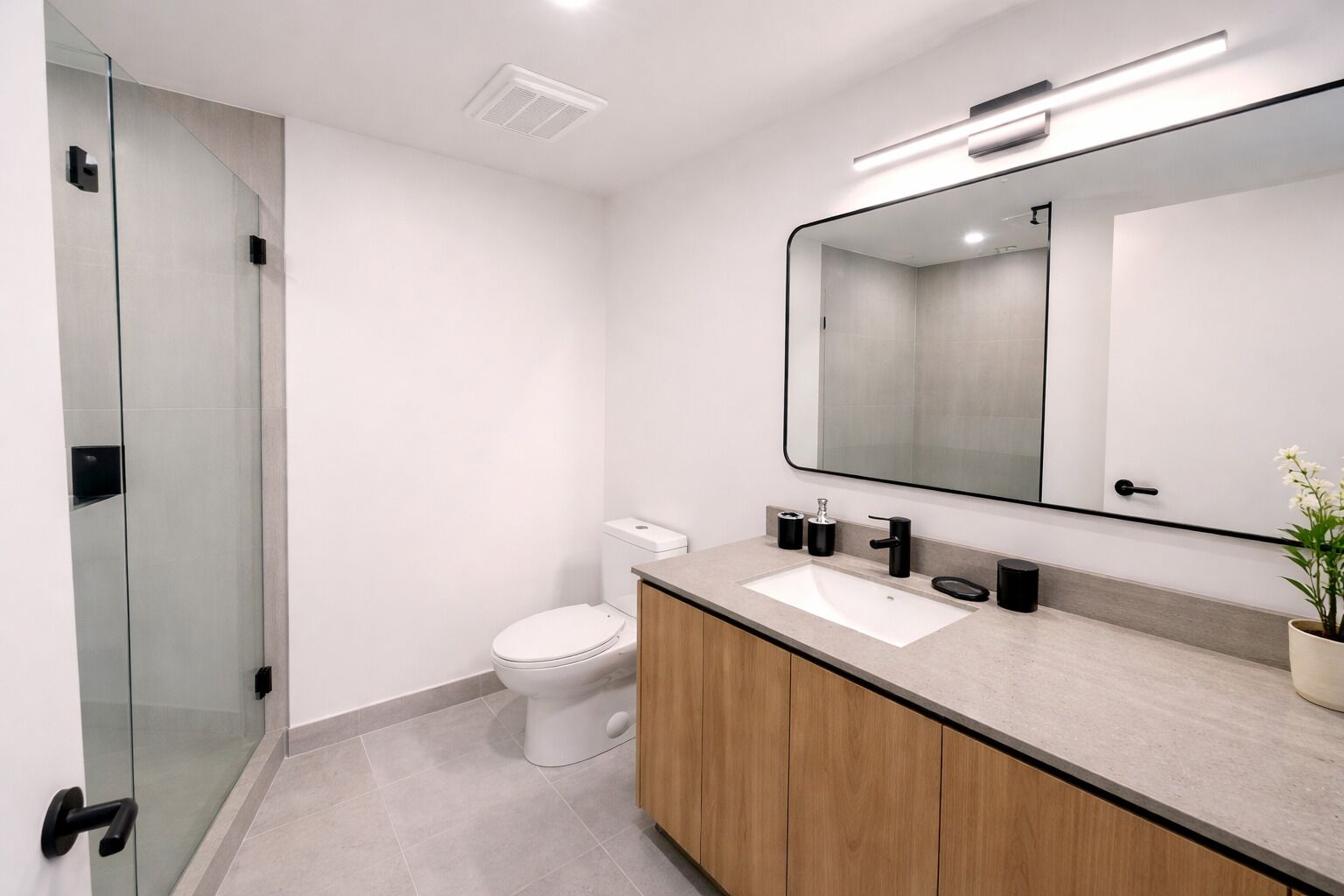 A modern bathroom with a glass shower enclosure, a white toilet, a wooden vanity with a sink, and a large mirror.
