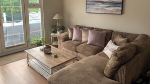 A living room with a beige sofa, a coffee table, and a lamp. There is a window with a view of a fence and greenery outside. - Spacious Lakeside Family Lodge in the Cotswolds | Sleeps 6 | Private Deck (Cirencester)