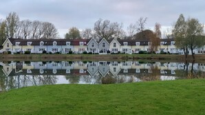 A row of traditional houses by a calm lake with a grassy foreground. - Spacious Lakeside Family Lodge in the Cotswolds | Sleeps 6 | Private Deck (Cirencester)