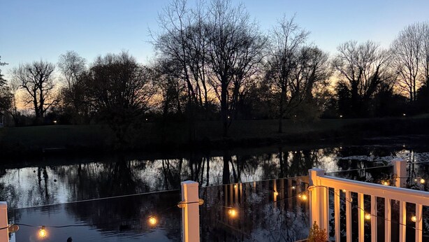 A wooden deck with a railing overlooking a calm body of water, surrounded by trees. - Spacious Lakeside Family Lodge in the Cotswolds | Sleeps 6 | Private Deck (Cirencester)