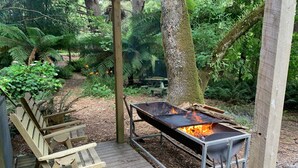 A wooden deck with a metal grill and fire pit, surrounded by lush greenery and a wooden bench.