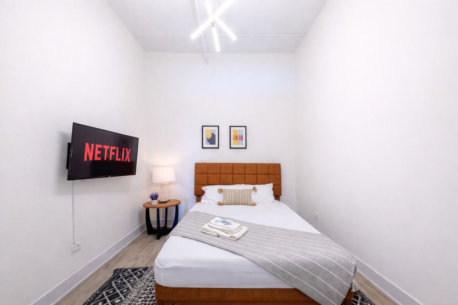 Habitación de hotel con cama, televisor montado en la pared, mesita de noche con lámpara y dos cuadros enmarcados.