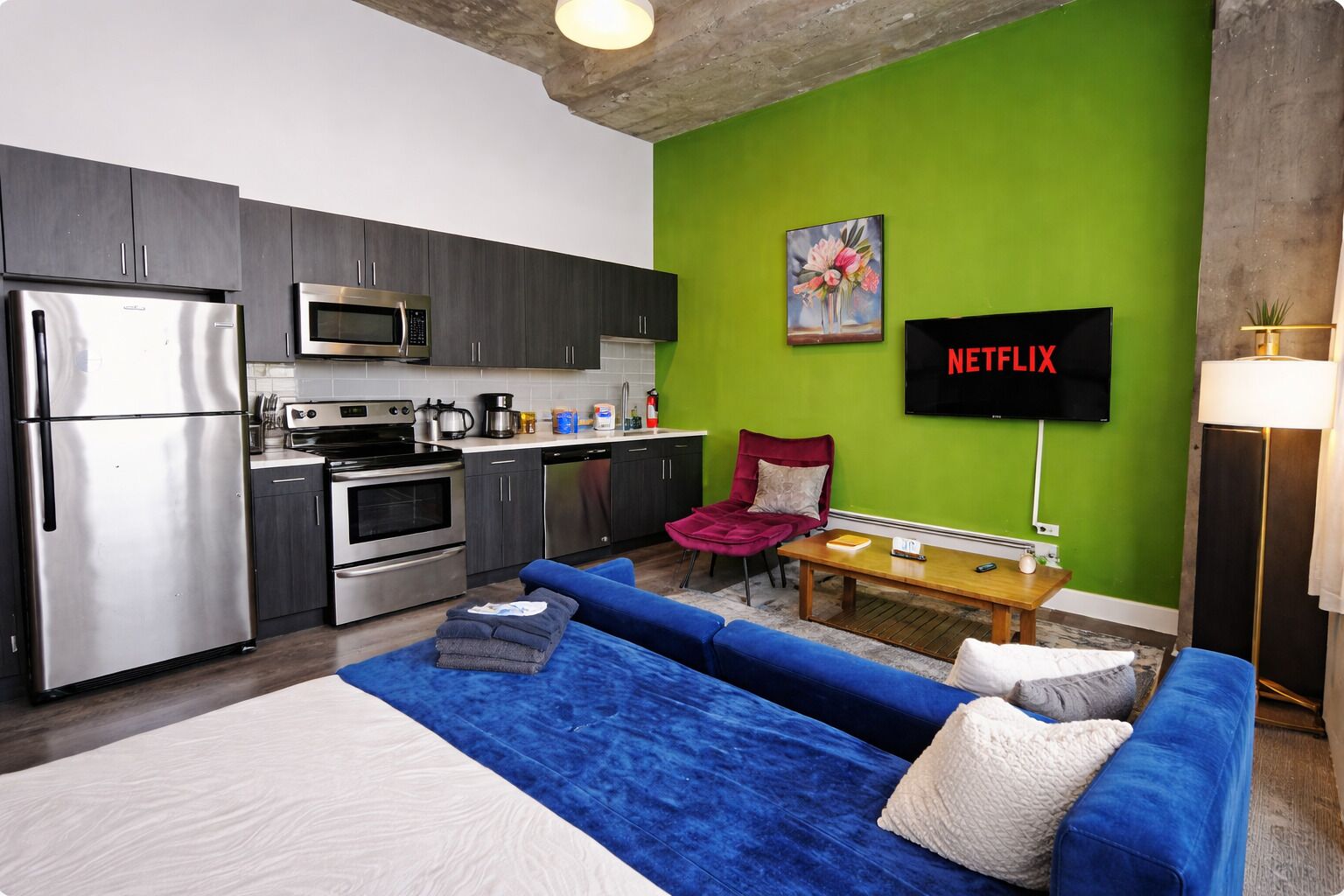 Ett kompakt kök med moderna vitvaror, en sittgrupp med en blå soffa och en röd fåtölj, och en väggmonterad TV som visar ”NETFLIX”.