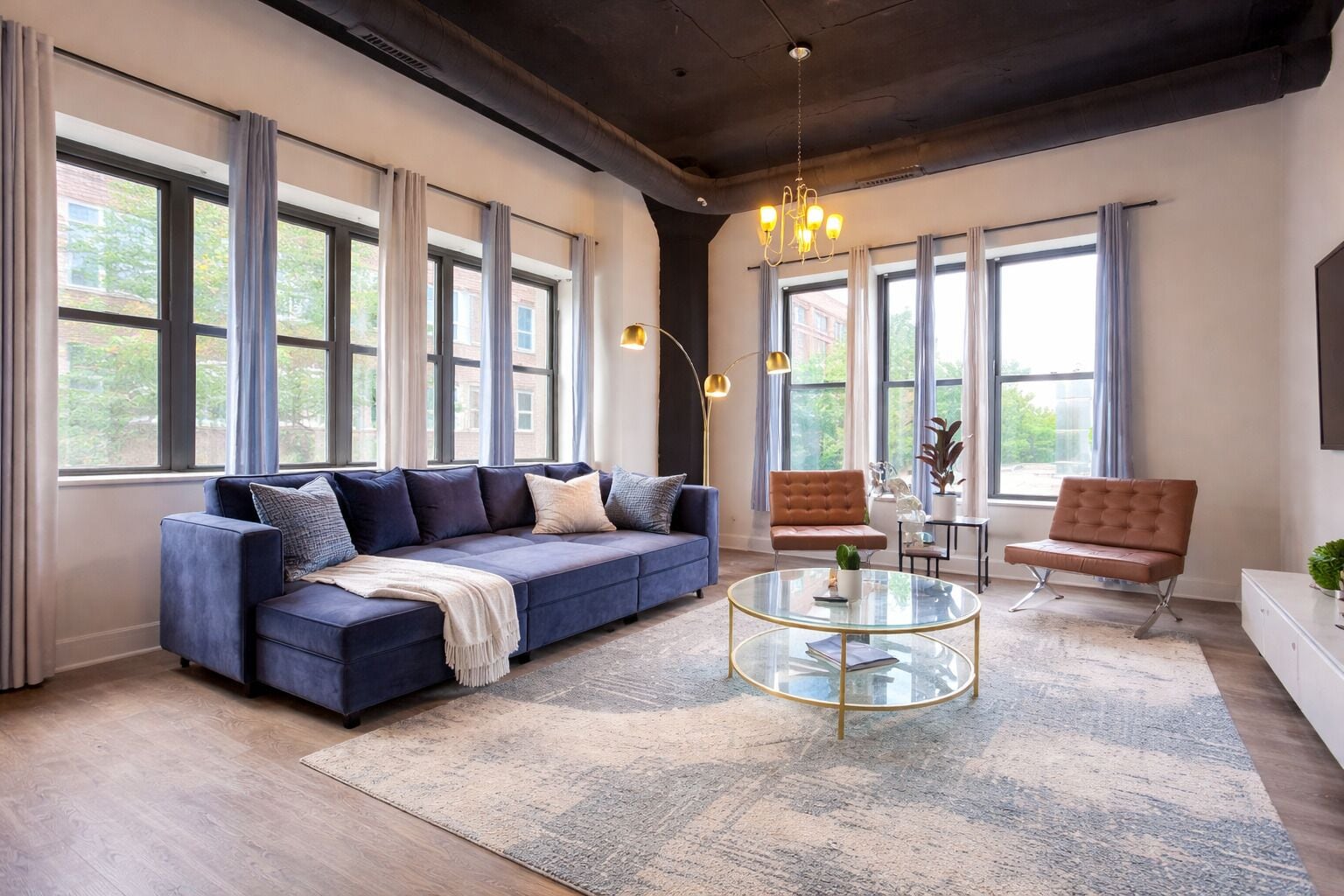 Ein modernes Wohnzimmer mit einem blauen Sofa, zwei Sesseln, einem Glastisch und großen Fenstern.