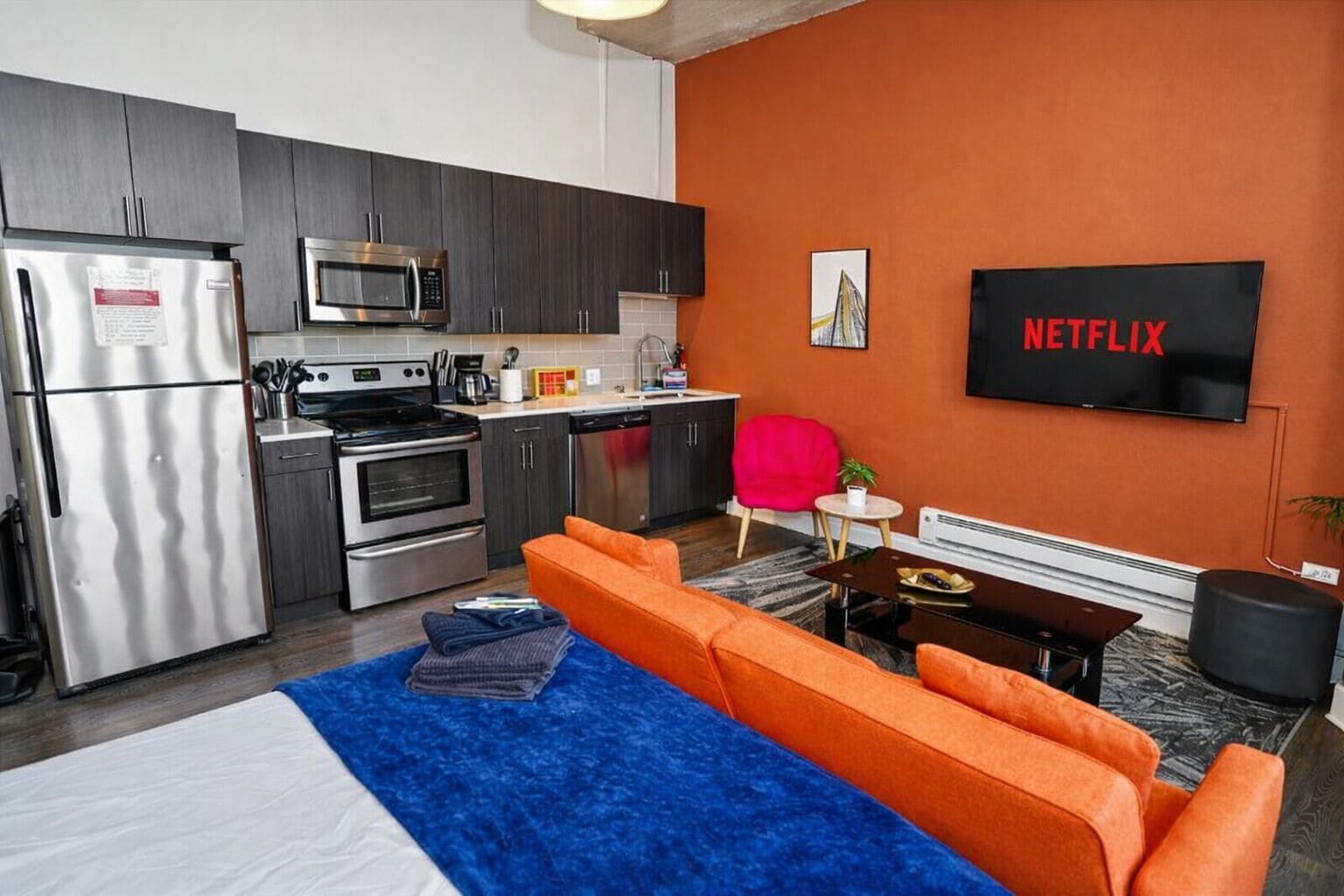 Habitación de hotel compacta con kitchenette, cama, sofá, televisión y una obra de arte colgada en la pared.