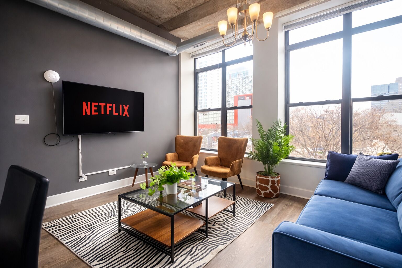 A modern living room with a large TV displaying 'NETFLIX,' a glass coffee table, two armchairs, a blue sofa, and a large window with a city view.