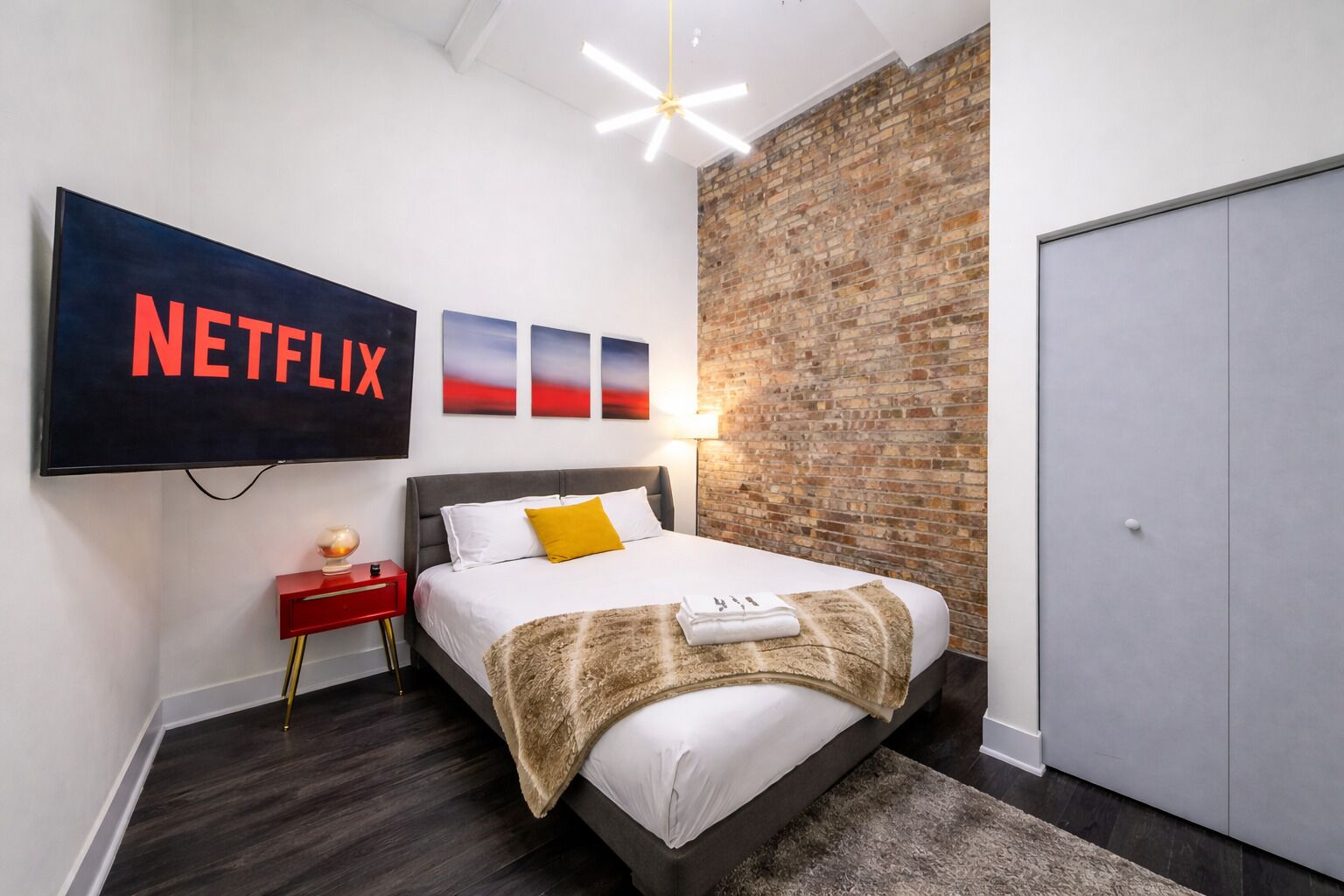 Une chambre à coucher moderne avec un grand lit, un téléviseur fixé au mur, un mur d’accent en briques et une armoire.
