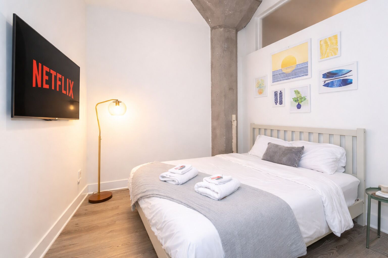 Ein ordentlich bezogenes Bett mit weißen Leinen, ein an der Wand befestigter Fernseher mit dem Netflix-Logo und eine Stehlampe.